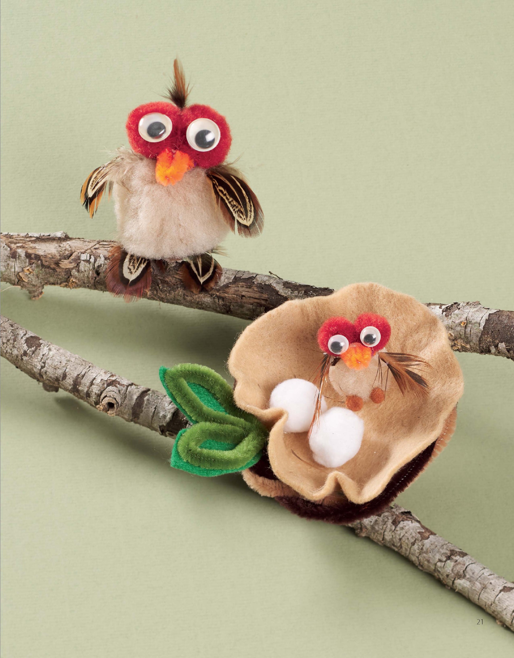 Two adorable owl crafts from Leisure Arts’ Pom-Pom Whimsy sit on tree branches, made with pom-poms and felt. One owl features bright red eyes, while a nest with two white eggs and felt leaves decorates the lower branch. Easy for beginners, these charming owls use chenille stems for wings and feet and are displayed against a soft green background.