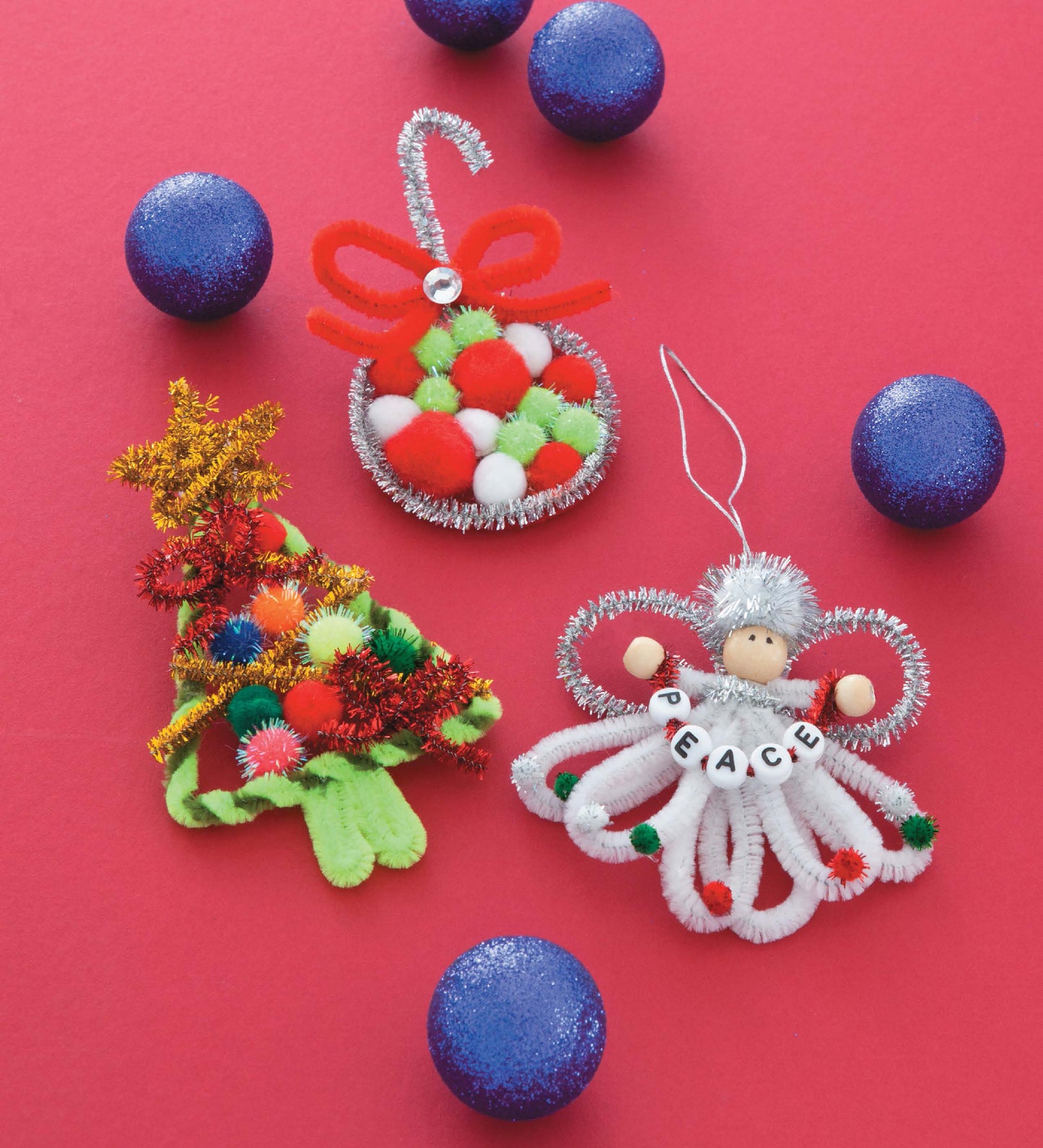 Three vibrant ornaments created from Pom-Pom Whimsy by Leisure Arts—a candy cane, Christmas tree, and angel adorned with PEACE beads—are crafted from chenille stems and surrounded by six sparkling blue pom-poms on a vivid red background. This cheerful arrangement is perfect for beginner crafters or as step-by-step project inspiration.