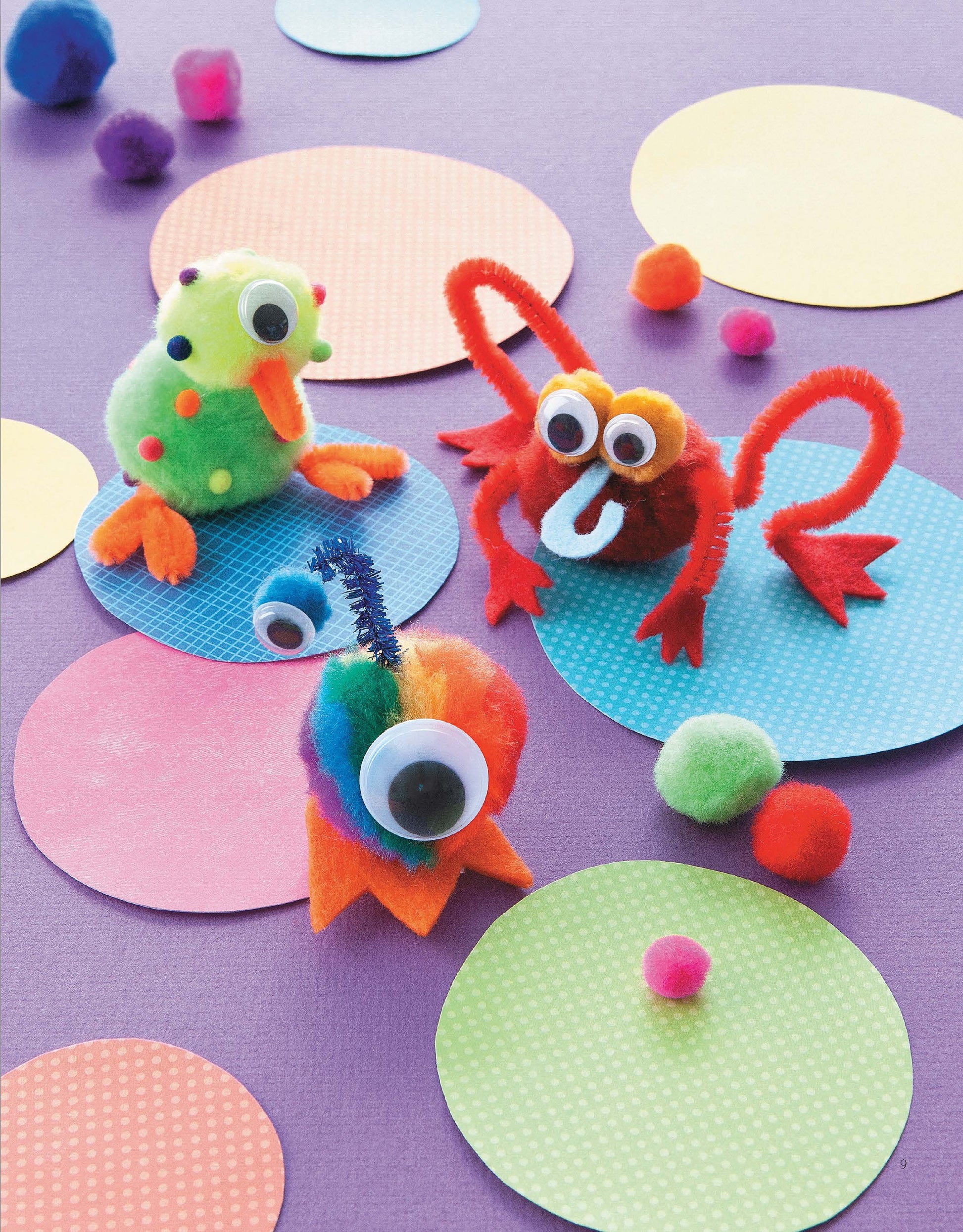 Three colorful, playful Pom-Pom Whimsy crafts from Leisure Arts, featuring googly eyes and chenille stem limbs, sit on dotted pastel circles against a purple background surrounded by small pom-poms—perfect for beginner crafters or step-by-step projects.
