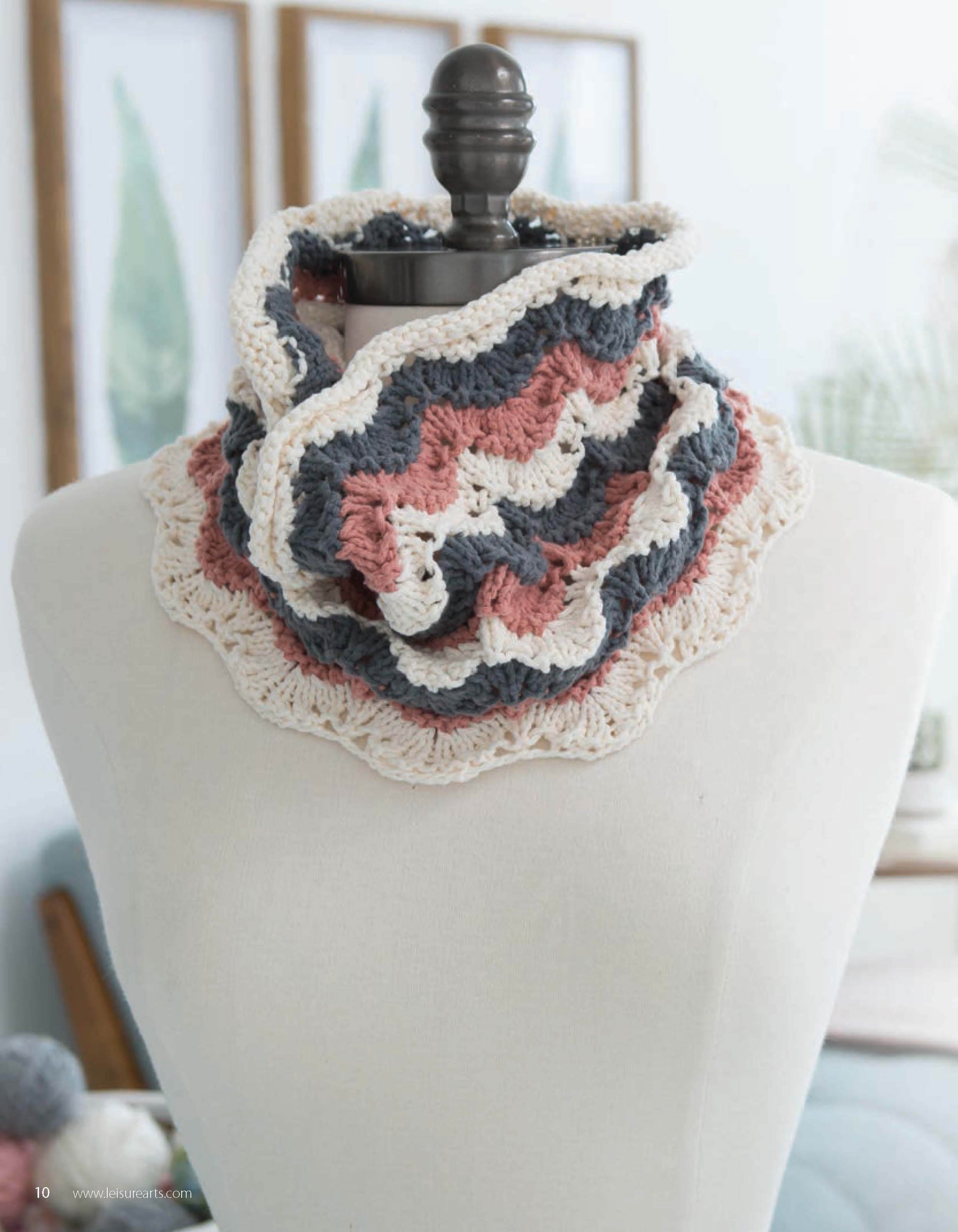 Lace Essentials by Leisure Arts, a crocheted ruffled cowl in cream, pink, and blue, is showcased on a mannequin bust with framed botanical prints and soft lighting creating a cozy backdrop.
