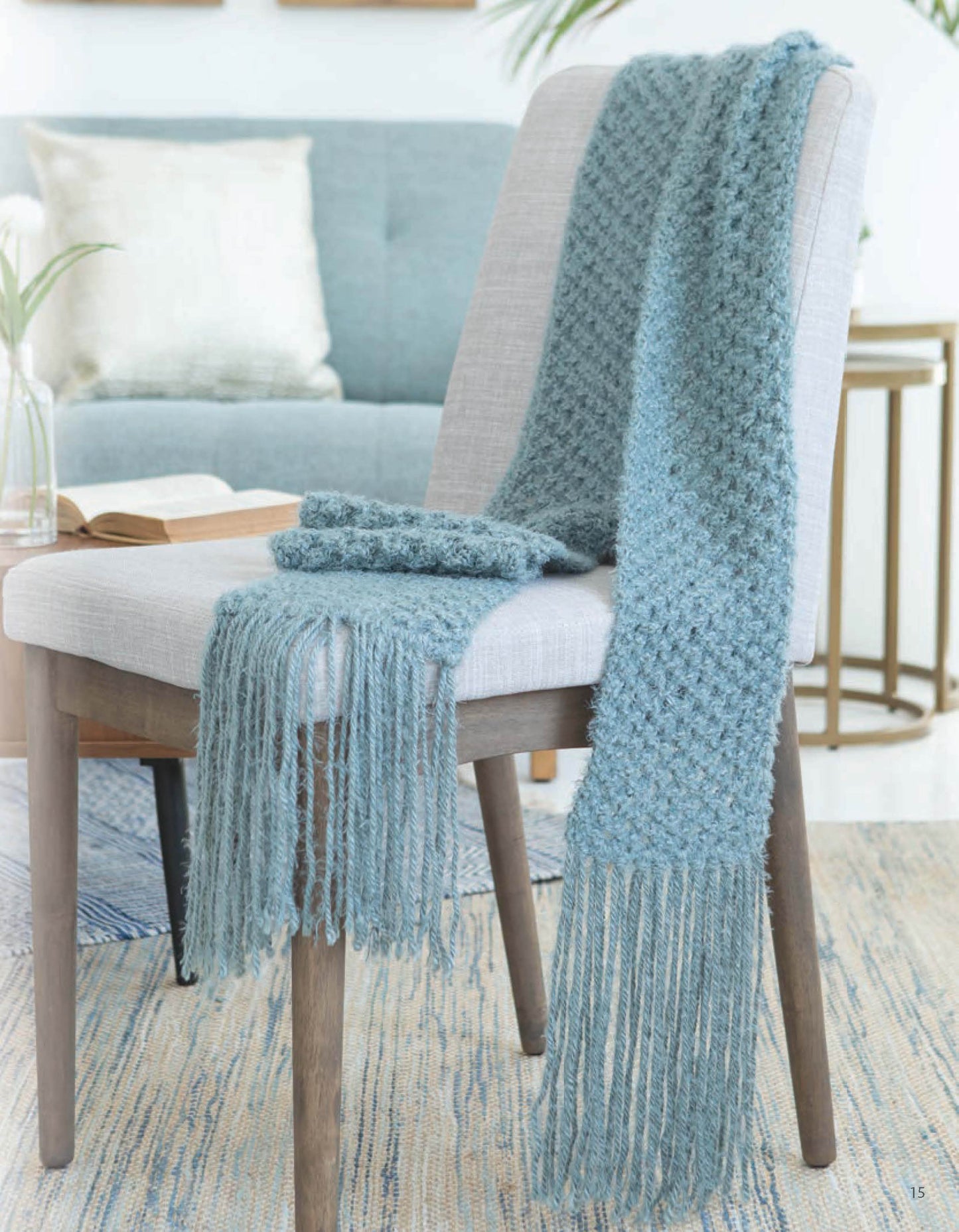The Leisure Arts Lace Essentials scarf in textured blue with long fringe is draped over a light gray chair. A matching pair of blue mittens rests on the seat, set against a cozy living room and sofa background.