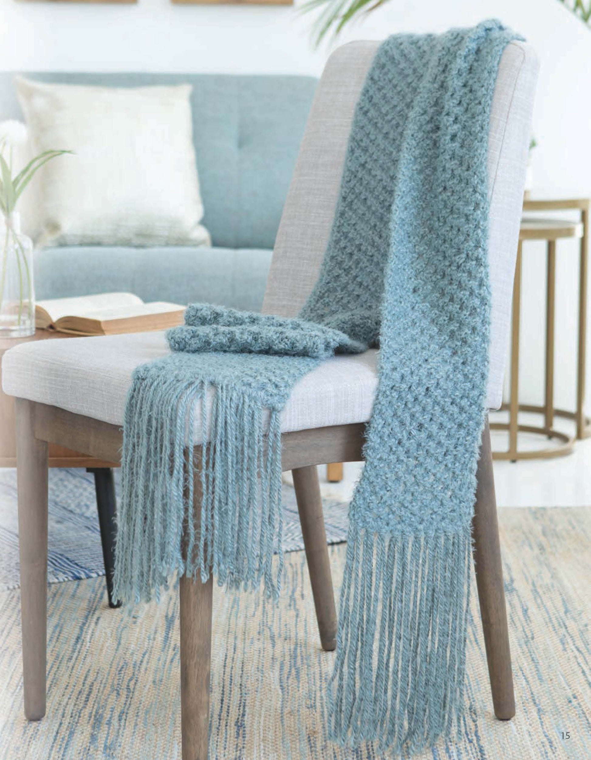 The Leisure Arts Lace Essentials scarf in textured blue with long fringe is draped over a light gray chair. A matching pair of blue mittens rests on the seat, set against a cozy living room and sofa background.