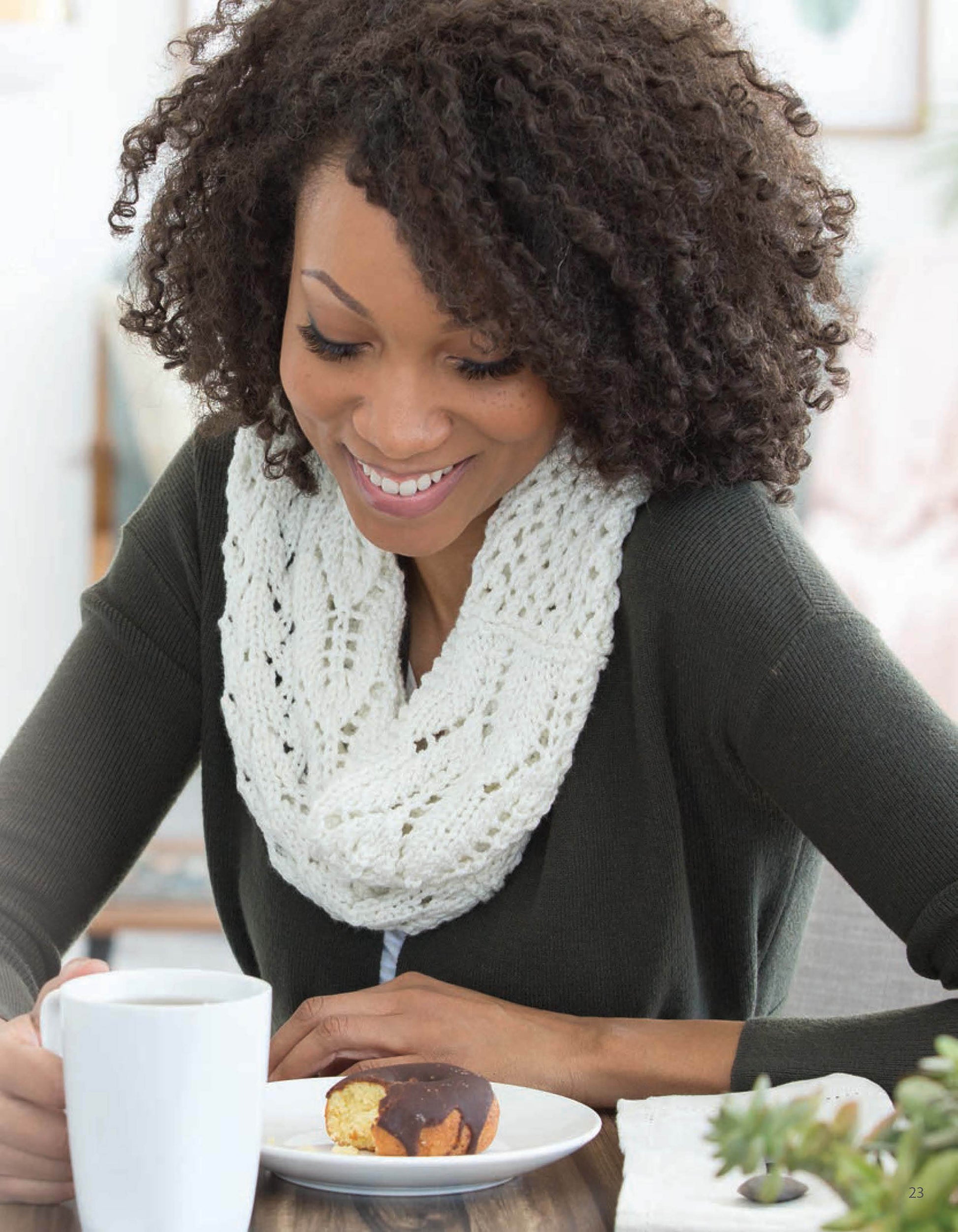 A woman with curly hair and a Leisure Arts Lace Essentials scarf smiles at a table, enjoying coffee and a chocolate-covered pastry.