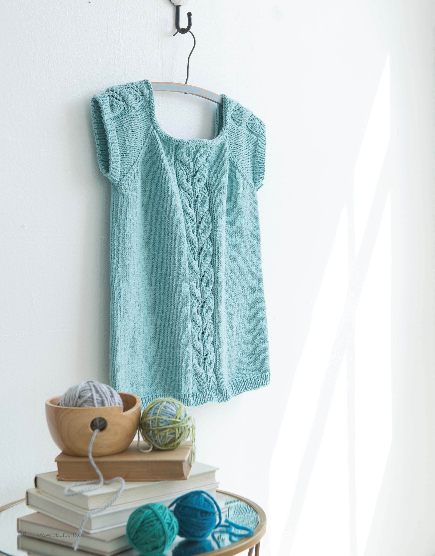The Lace Tops & Vests by Leisure Arts is a knitted teal sleeveless top with lace cable detail, shown on a hanger above a table with yarn balls and books—perfect inspiration for knitting or exploring new lace patterns.