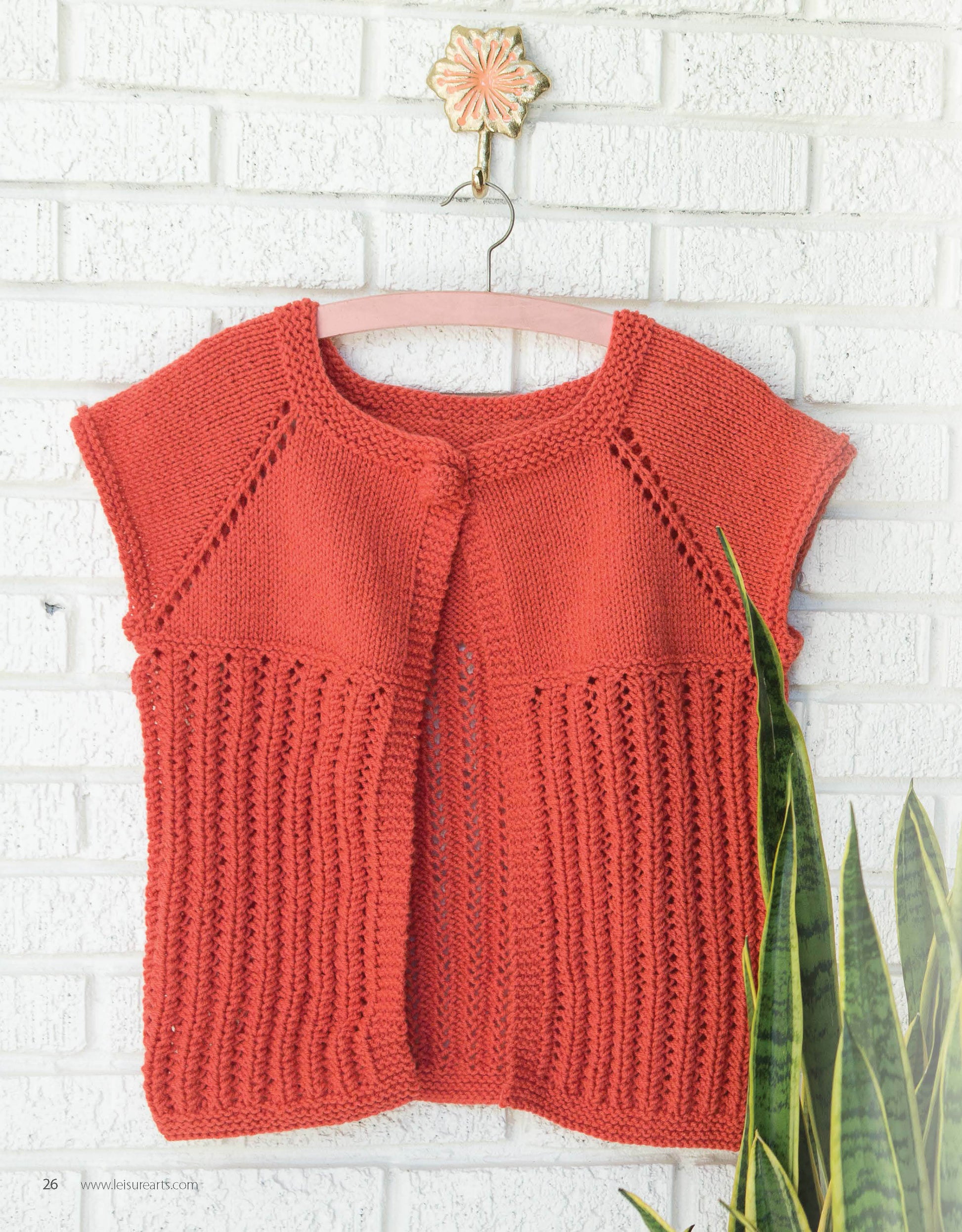 The Leisure Arts Lace Tops & Vests, a hand-knitted orange cardigan with short sleeves and ribbed front panels, hangs on a wooden hanger by a white brick wall, with a green snake plant partially visible in the corner.