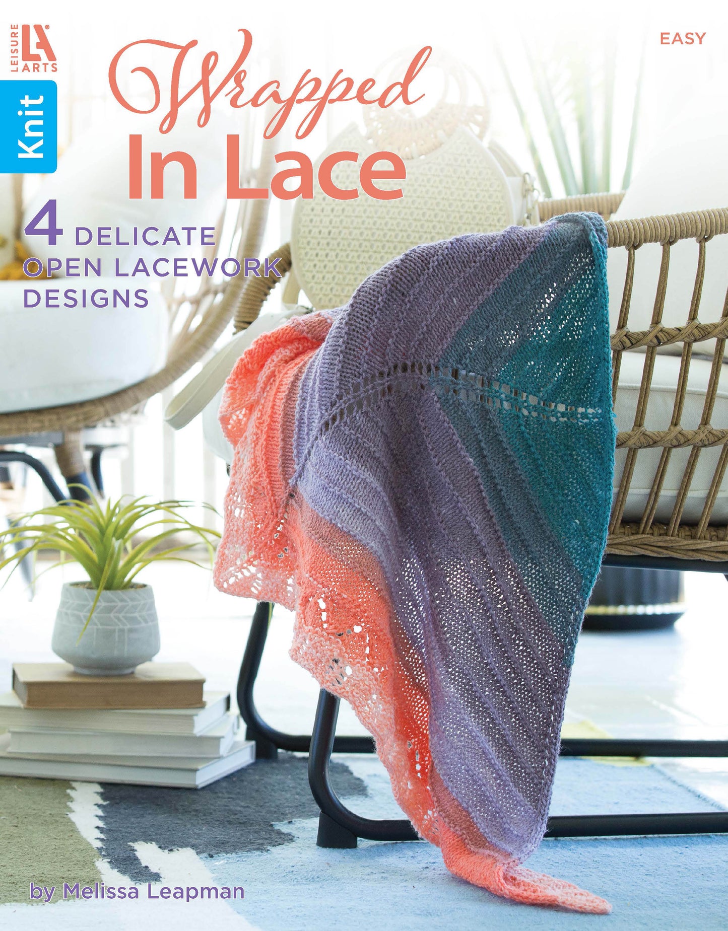 A vibrant lace knit blanket in peach, teal, and lavender is draped over a black chair on the cover of "Wrapped in Lace" by Leisure Arts, showcasing beautiful lace knitting patterns.