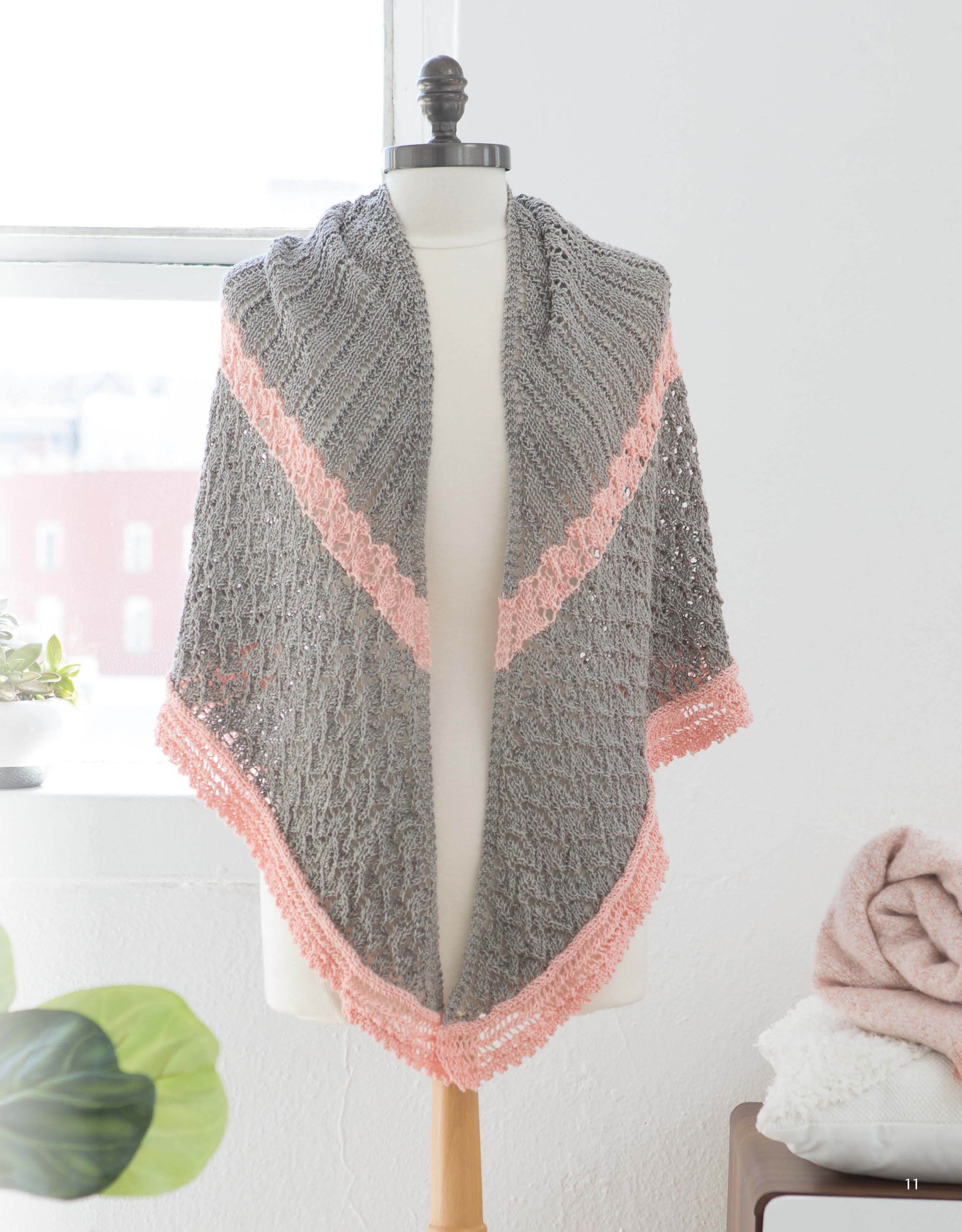 A mannequin showcases the Leisure Arts "Wrapped in Lace" shawl, gray with pink trim and delicate lace patterns, draped over its shoulders in a bright room with a window, plant, and folded blankets in the background.