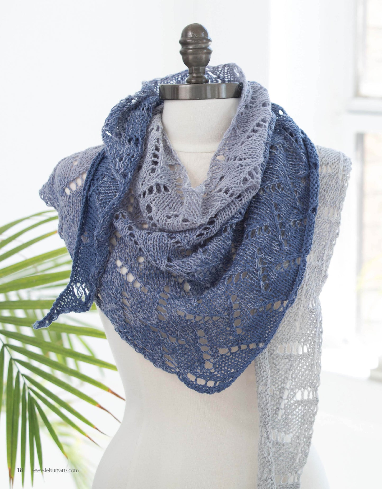 The mannequin models Leisure Arts' "Wrapped in Lace," a triangular lace-knit scarf with light gray to deep blue gradient. A green plant and sunlight through the window create a bright background.