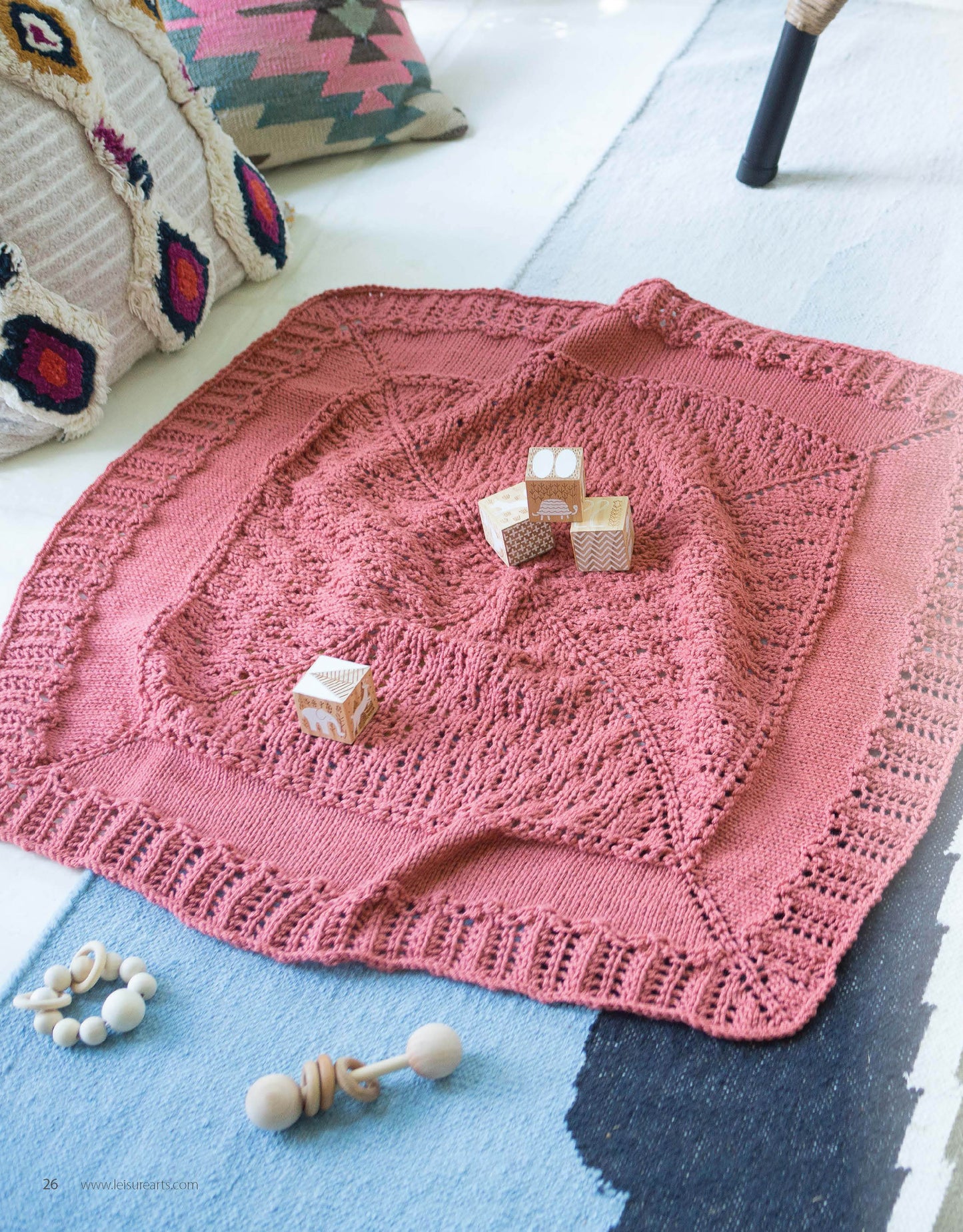 The Wrapped in Lace baby blanket from Leisure Arts, with its delicate lacy border, is displayed on a light blue rug. Wooden toy blocks, rattles, and patterned cushions add charm to this cozy and inviting knitting project scene.