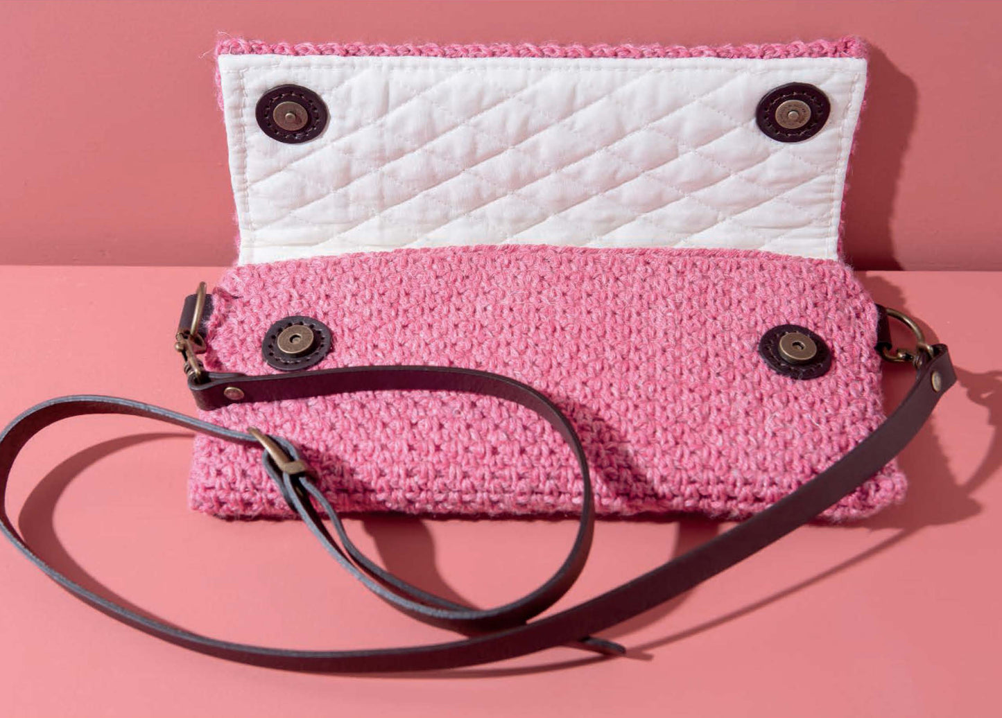 Inspired by patterns from Leisure Arts’ Crochet Handcrafted Handbags Book 1, this pink crocheted bag features a quilted white lining, four magnetic snaps, and a dark brown leather shoulder strap. Shown open on a pink surface.