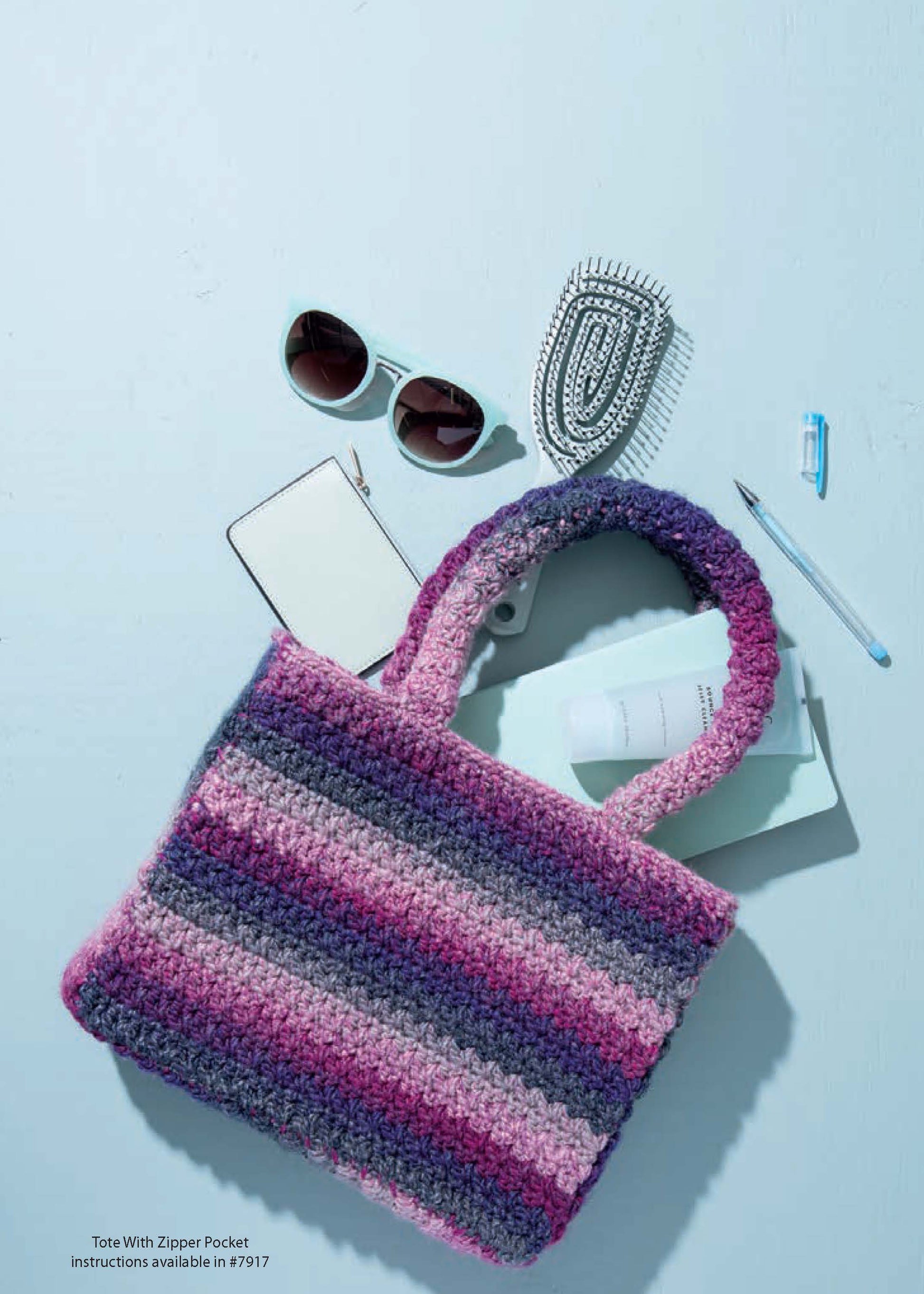 A pink and purple striped crocheted tote bag rests on a light blue surface with round sunglasses, a notebook, the Leisure Arts "Crochet Handcrafted Handbags Book 2," a pen, a silver beaded purse, and lip balm nearby.