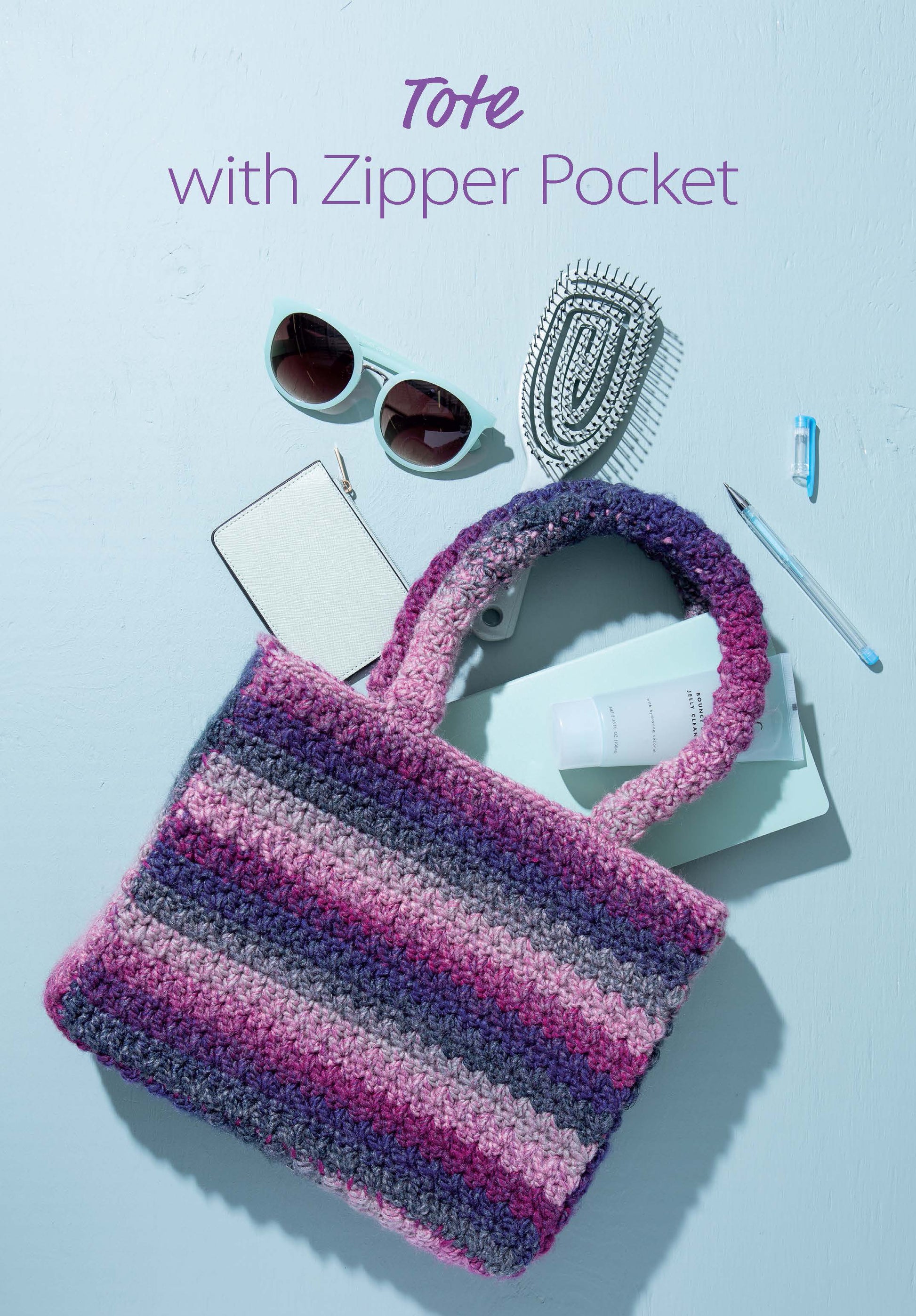 The image shows items on a light blue surface with a crocheted purple and pink striped tote from Leisure Arts' "Crochet Handcrafted Handbags Book 3" digital download. Text above the tote reads, "Tote with Zipper Pocket.