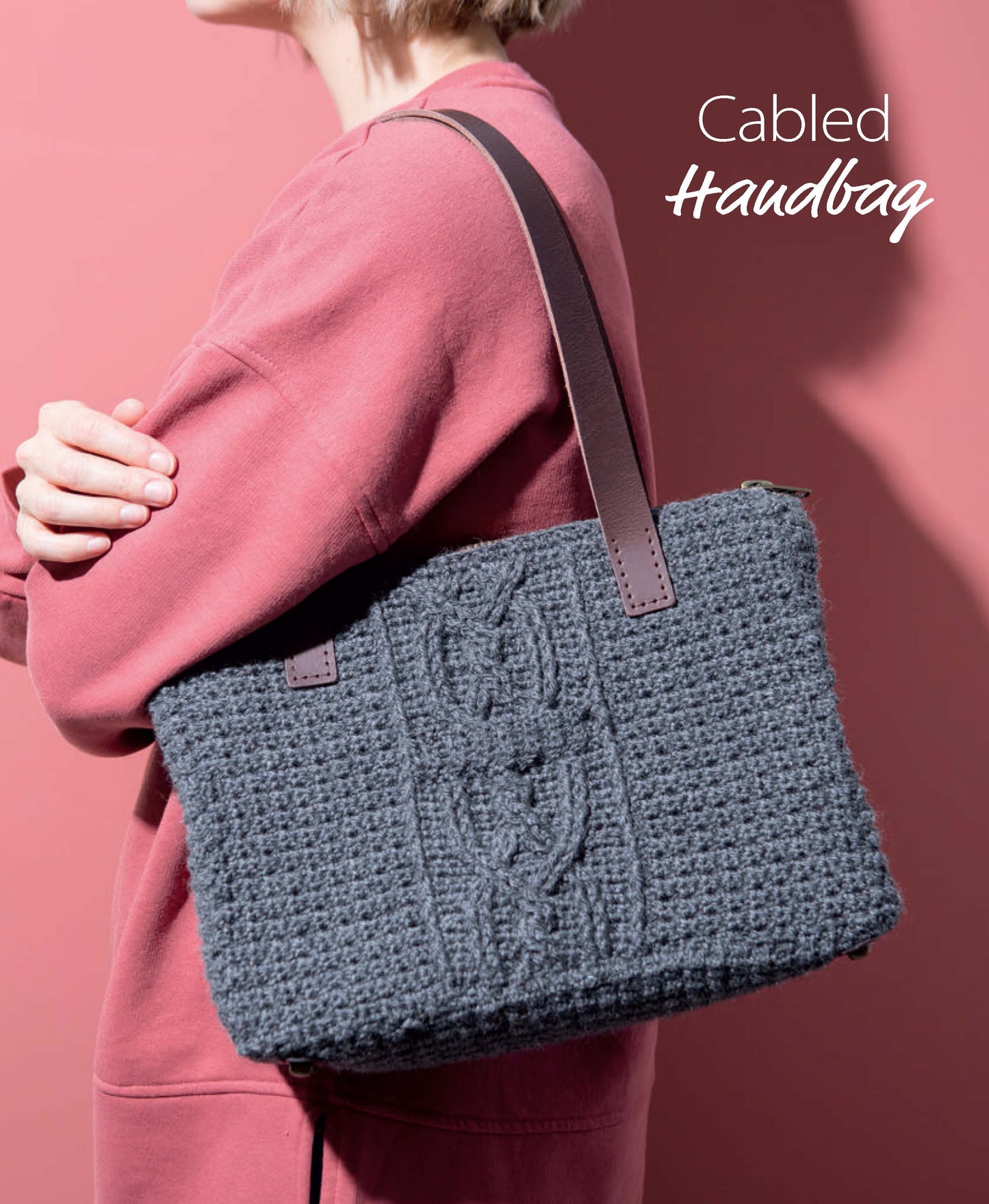 Against a pink background, someone in a pink sweater holds a textured dark gray crocheted handbag with leather straps. Find inspiration in Leisure Arts’ Crochet Handcrafted Handbags Book 4. "Cabled Handbag" text is shown in the upper right corner.