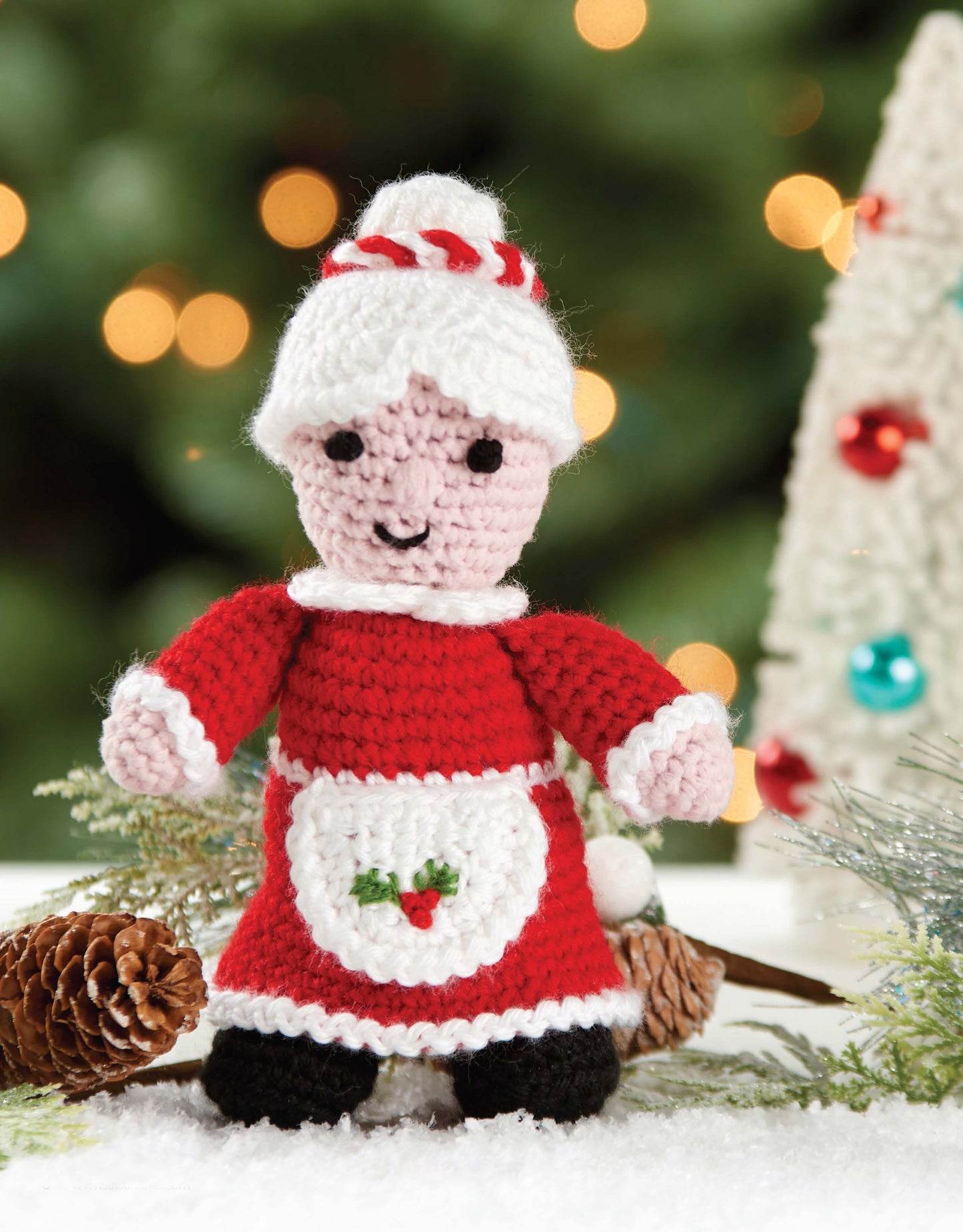 The Leisure Arts Christmas Friends set features 4 adorable crocheted Christmas characters, including Mrs. Claus, displayed on snow with pinecones, greenery, and a festive tree in the background.