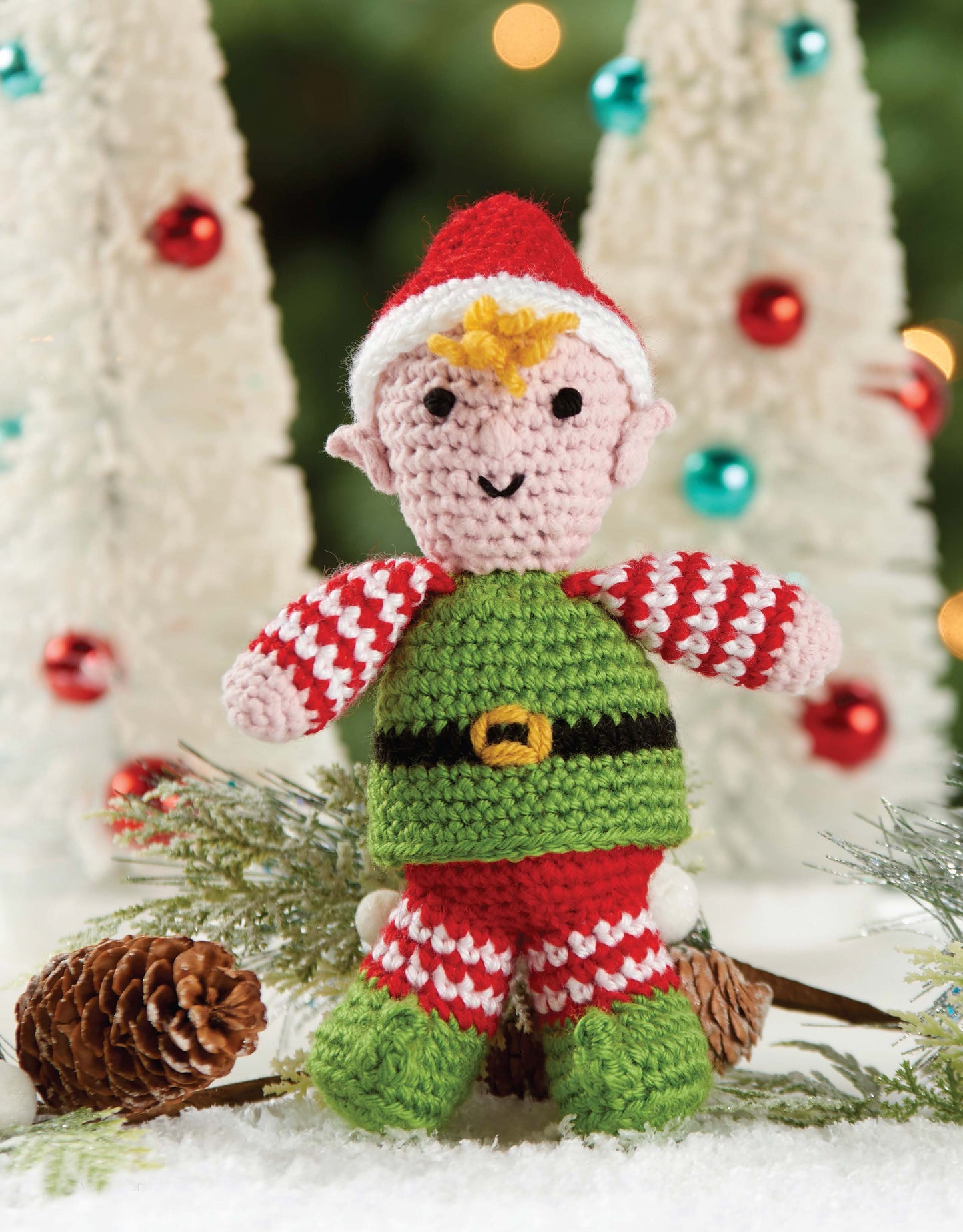 Leisure Arts' Christmas Friends—4 Adorable Christmas Characters features a crocheted elf in a red hat, striped sleeves, green outfit with yellow belt, posed among pinecones and snowy greenery with white Christmas trees behind.